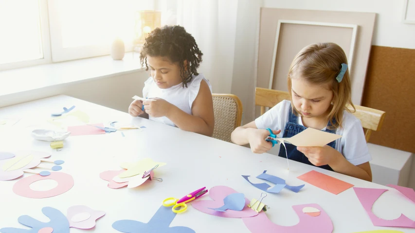 two little girls sitting at a table making crafts, by Julian Allen, paper cutouts of plain colors, promo image, each having six wings, natural morning light