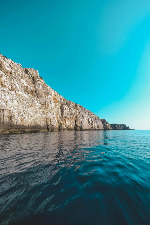 a body of water with a cliff in the background, a picture, by Carlo Martini, unsplash, romanticism, clear blue sky, maritime, slide show, teal color graded