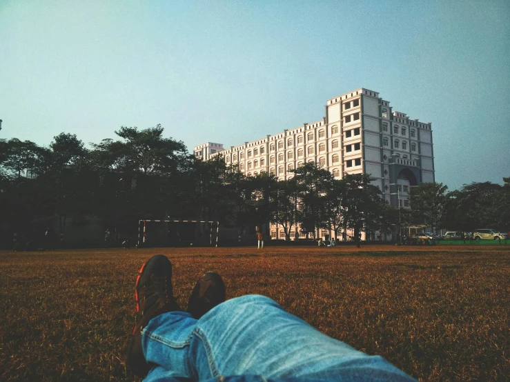 a person laying in a field with a building in the background, unsplash contest winner, bengal school of art, with stalinist style highrise, photo taken on fujifilm superia, chill time. good view, instagram photo amazing view