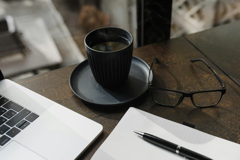 a laptop computer sitting on top of a wooden table next to a cup of coffee, by Carey Morris, pexels contest winner, round black glasses, fountain pen, dark black porcelain skin, paper cup