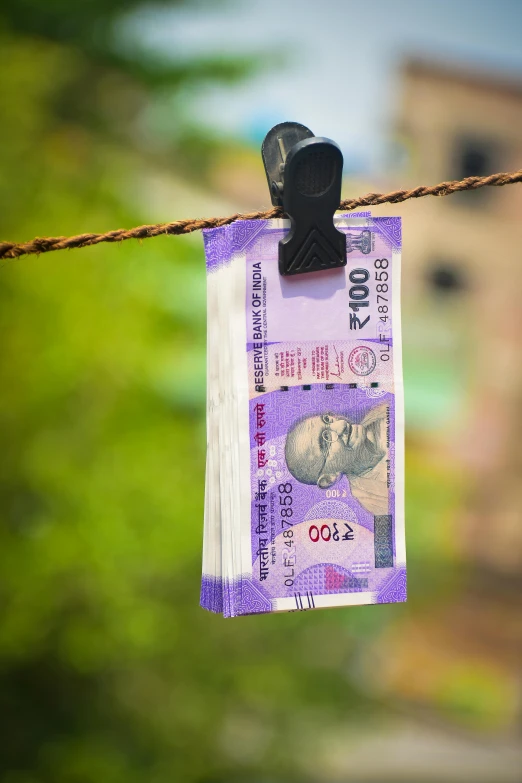 a note hanging on a clothes line with a building in the background, a portrait, by Jitish Kallat, pexels contest winner, banknote, screensaver, hindu, leaked