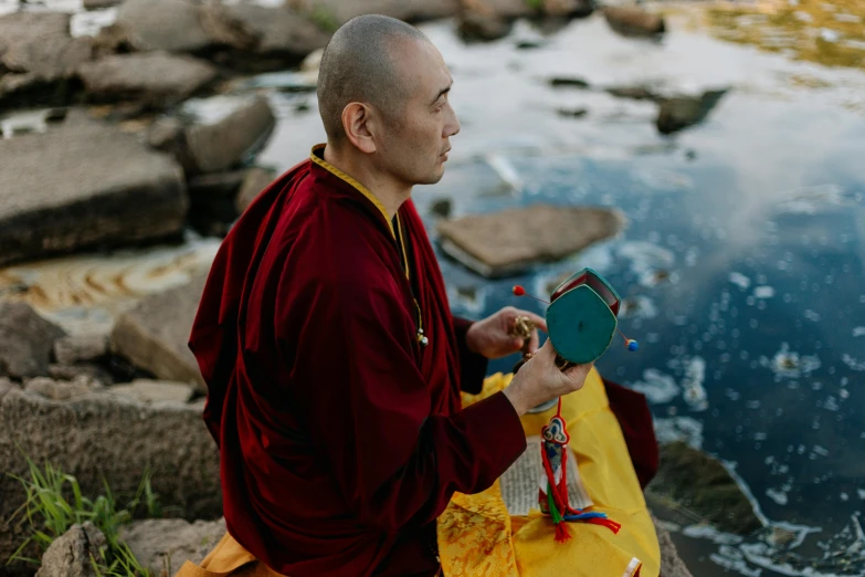 a man sitting on a rock next to a body of water, wearing red robes, holding a gold bag, focused light, avatar image