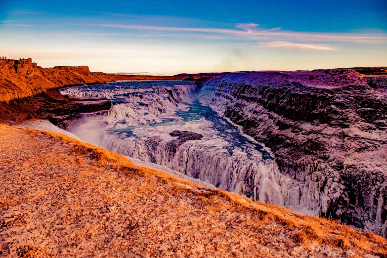 a man standing on top of a cliff next to a waterfall, by Jesper Knudsen, pexels contest winner, hurufiyya, sunset glow, panoramic, crater, indigo