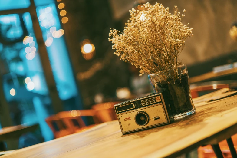 a camera sitting on top of a wooden table, inspired by Elsa Bleda, unsplash contest winner, inside a french cafe, flowers around, retro stylised, camera flash