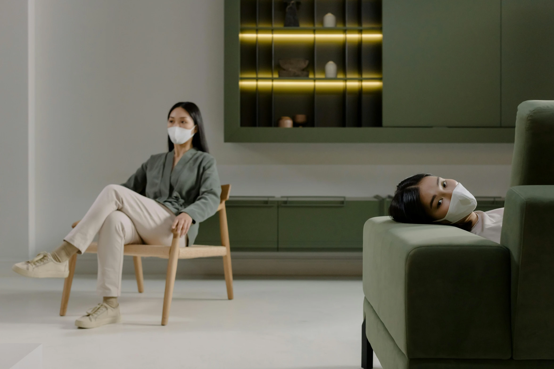 a woman sitting in a chair with a face mask on, inspired by Fei Danxu, trending on pexels, video art, two people, minimalist furniture, cinematic wide shot, green facemask