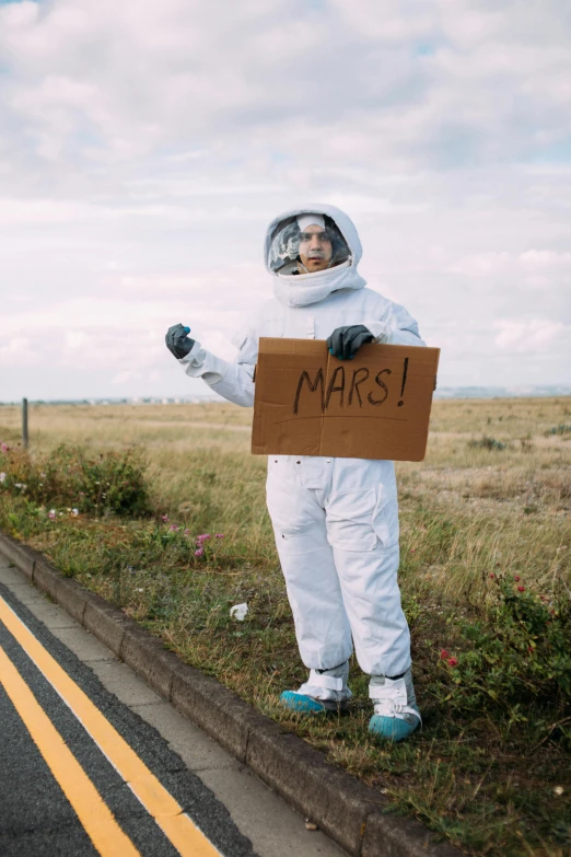 a person in a space suit holding a sign, pexels contest winner, happening, road trip, marc, non-binary, on ground