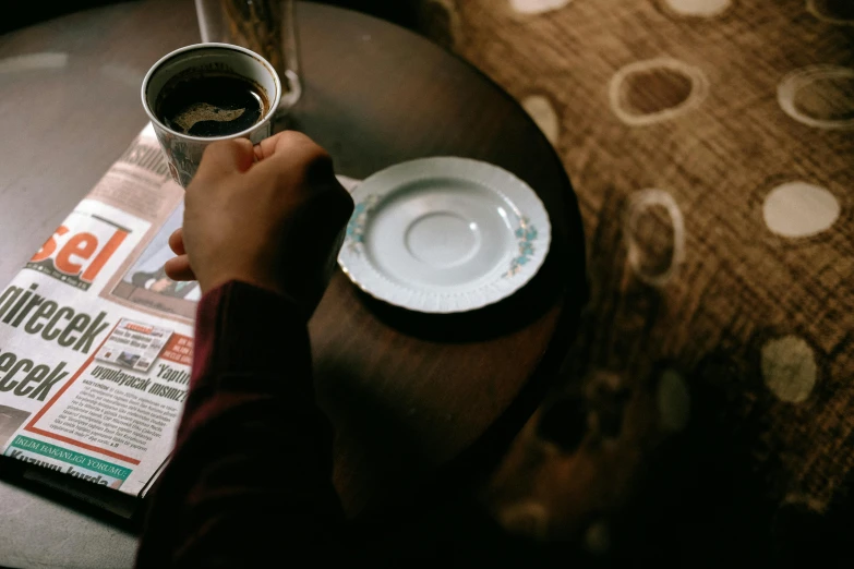 a person sitting at a table with a cup of coffee, by Elsa Bleda, pexels contest winner, newspaper, served on a plate, 15081959 21121991 01012000 4k, on a coffee table