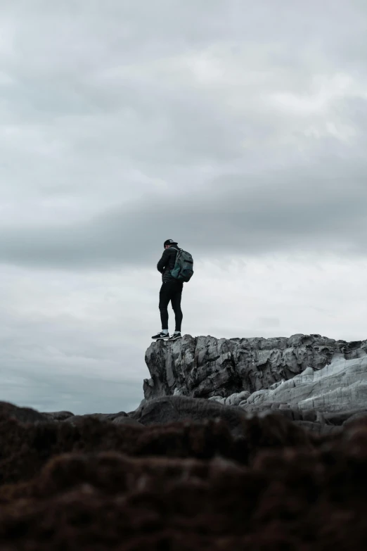 a person standing on top of a rocky hill, grey skies, with a backpack, trending photo, full body dramatic profile