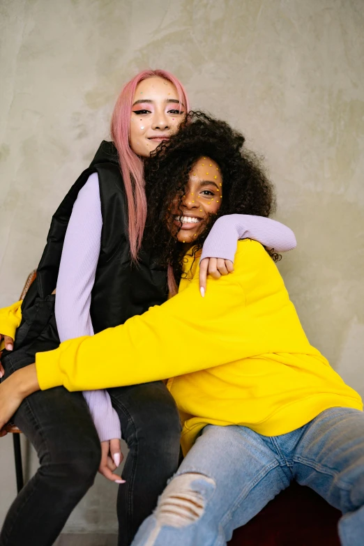 two women sitting next to each other on a chair, trending on pexels, antipodeans, wearing a yellow hoodie, curly pink hair, hugging, mix of ethnicities and genders