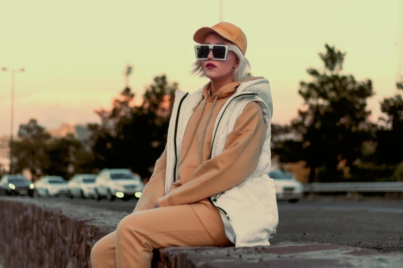 a woman sitting on top of a cement wall, inspired by Elsa Bleda, trending on pexels, realism, beige hoodie, sunglasses, yellow overall, wearing a track suit