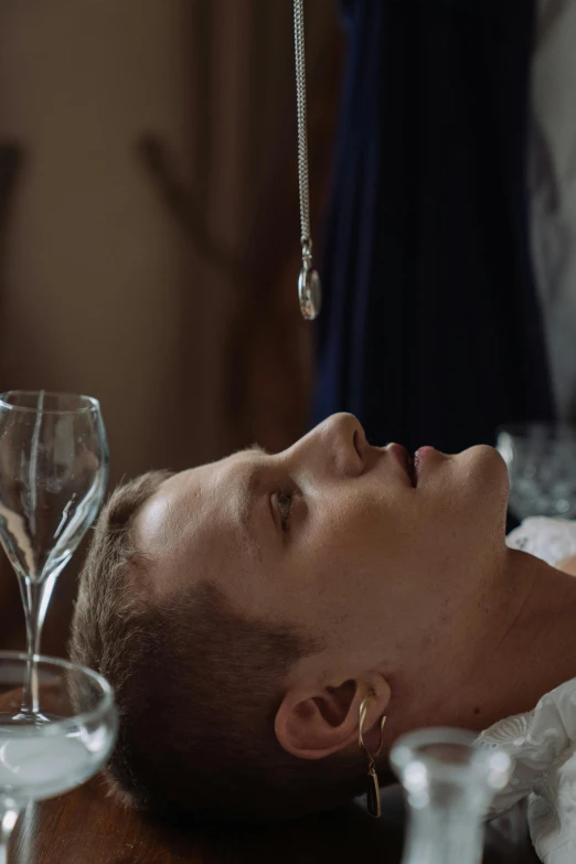 a man laying on top of a table next to wine glasses, inspired by Nikolaj Abraham Abildgaard, renaissance, huge earrings and queer make up, movie still frame, sacrament, intravenous drip