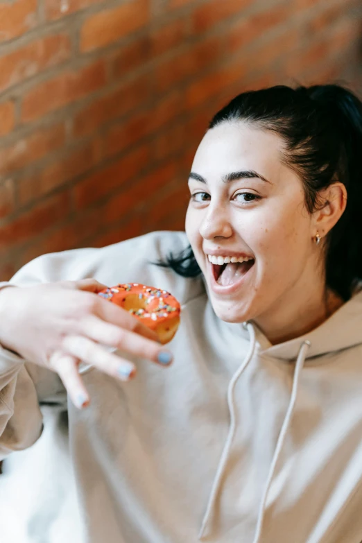 a woman eating a donut in front of a brick wall, pexels contest winner, antipodeans, wearing a hoodie and sweatpants, she is smiling and excited, charli xcx, people inside eating meals