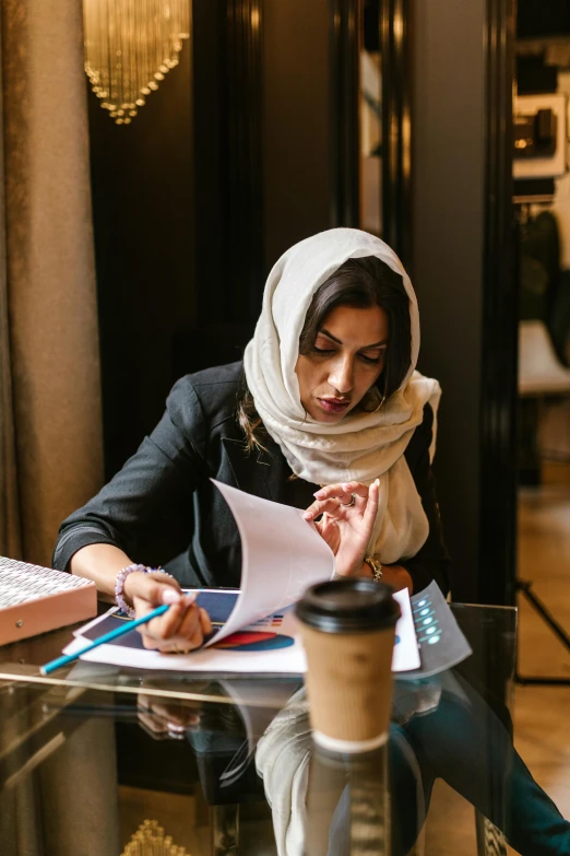 a woman sitting at a table with a cup of coffee, by Maryam Hashemi, pexels contest winner, academic art, working in an office, wearing a head scarf, writing on a clipboard, wearing business casual dress