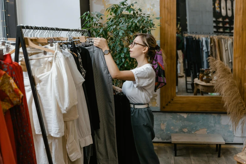 a woman standing in front of a rack of clothes, trending on pexels, happening, inspect in inventory image, white shirt and grey skirt, presenting wares, foliage clothing