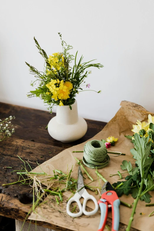 a bunch of flowers sitting on top of a wooden table, a still life, unsplash, arts and crafts movement, yellow and greens, flower shop scene, white vase, wrapped in flowers and wired