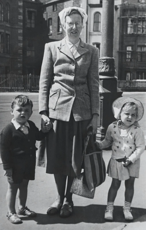 a black and white photo of a woman and two children, standing in a city center, post war style, in scotland, straw hat and overcoat