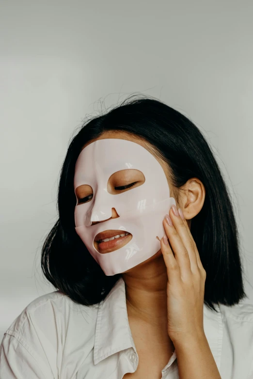 a woman putting a sheet mask on her face, trending on pexels, south east asian with round face, set against a white background, silicone skin, on a gray background