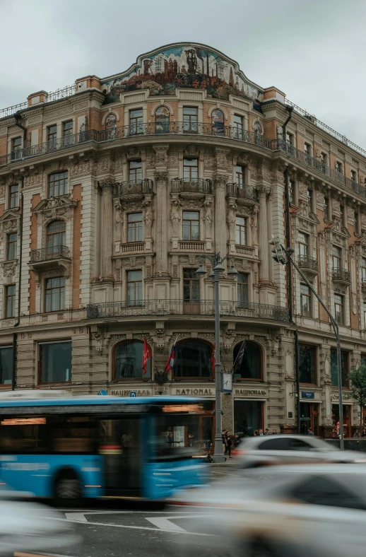 a blue bus driving down a street next to a tall building, by andrei riabovitchev, pexels contest winner, art nouveau, russian neoclassicism, panorama, nice slight overcast weather, gif
