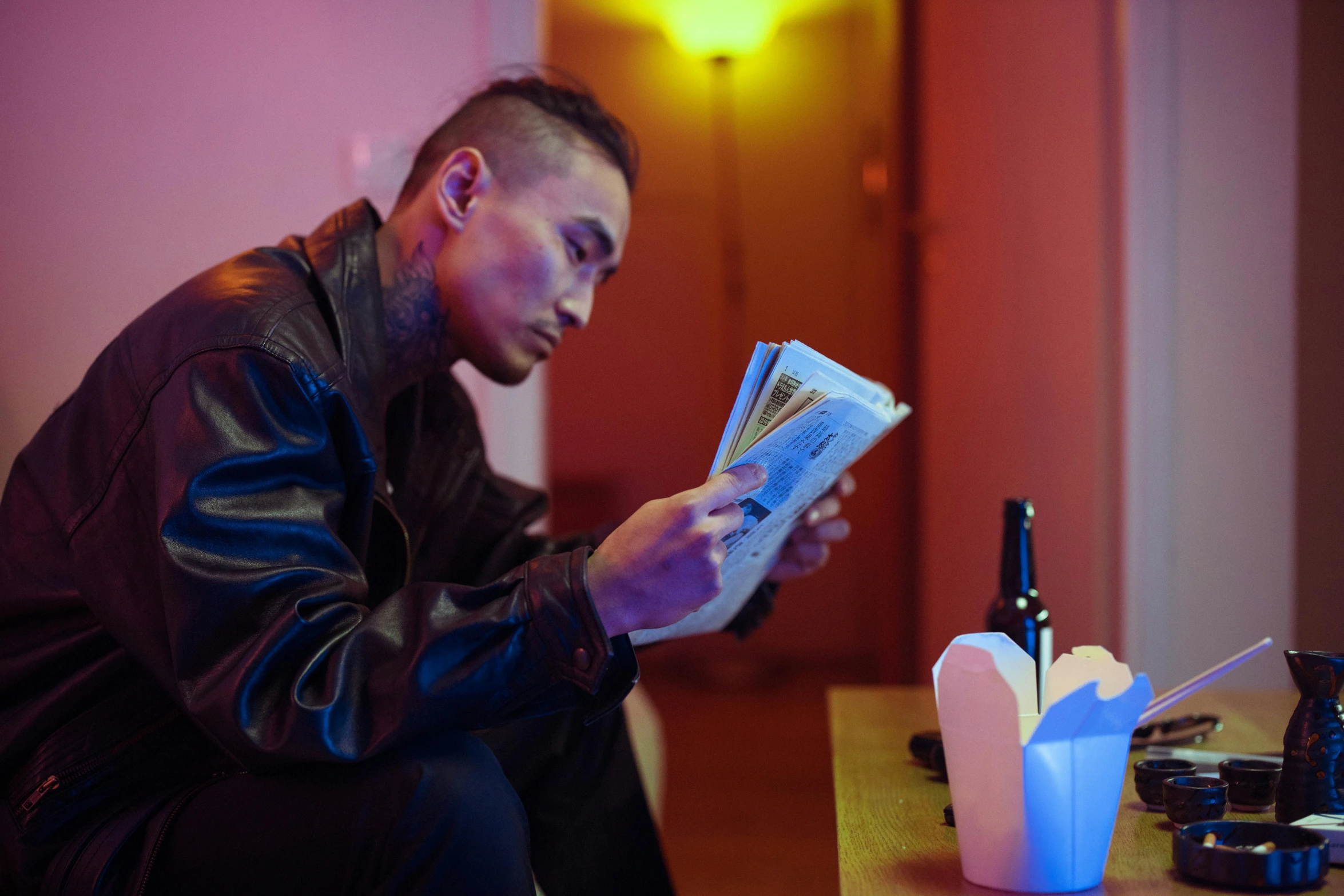 a man sitting at a table reading a book, inspired by Liam Wong, wearing cyberpunk leather jacket, reading for a party, profile image, cash on a sidetable