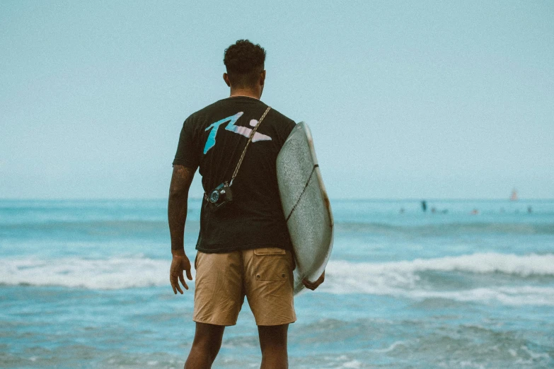 a man standing on a beach holding a surfboard, pexels contest winner, afrofuturism, wearing a black tshirt, back turned, wearing shorts and t shirt, avatar image