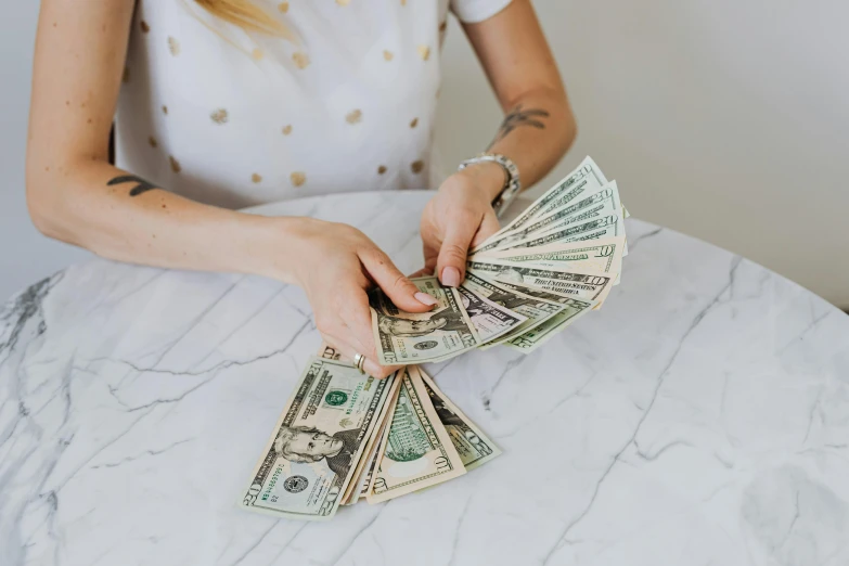 a woman sitting at a table holding a stack of money, by Emma Andijewska, pexels contest winner, fan favorite, on a marble pedestal, 🚀🌈🤩, society 6