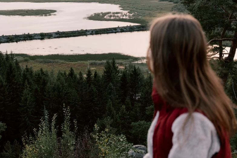a woman standing on top of a lush green hillside, by Emma Andijewska, pexels contest winner, gazing at the water, late summer evening, detailed lake in background, looking out at a red ocean