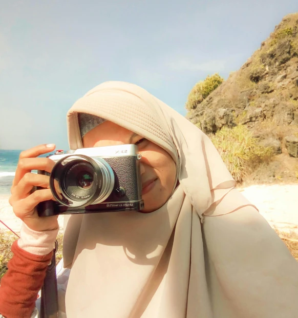 a woman taking a picture with a camera, inspired by Nazmi Ziya Güran, instagram, hijab, beach, low colour, cheeks
