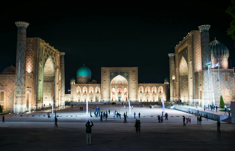 a group of people standing in front of a building at night, hurufiyya, islamic architecture, square, distant photo, ash thorp khyzyl saleem