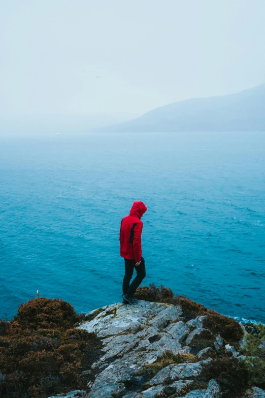 a person standing on top of a rock near the ocean, wearing a red hoodie, in the rain, pondering, overlooking