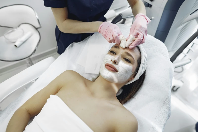 a woman getting a facial mask at a beauty salon, a photo, by Julian Allen, shutterstock, massurrealism, nurse, square facial structure, youtube thumbnail, petite girl