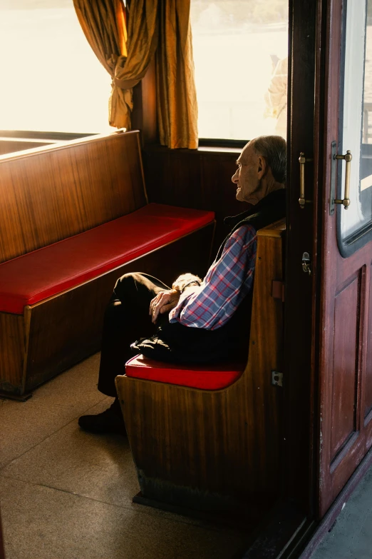 a man sitting on a bench in a train car, by Peter Churcher, winter sun, on a boat, colour photograph, frail