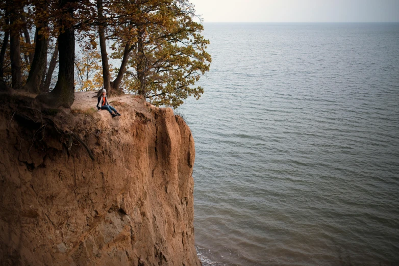 a man sitting on top of a cliff next to the ocean, by Jesper Knudsen, trees and cliffs, ukraine. photography, thumbnail, multiple stories