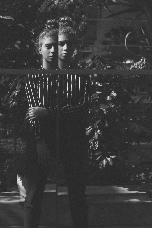 a black and white photo of a woman standing in front of a tree, a black and white photo, tumblr, realism, wearing stripe shirt, next to a plant, medium format. soft light, behind bars