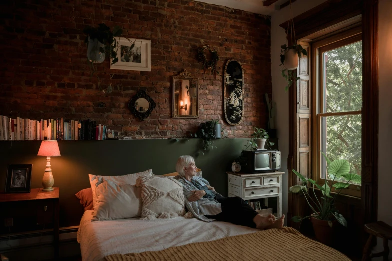 a woman sitting on top of a bed next to a window, inspired by Elsa Bleda, unsplash contest winner, old brick walls, elderly woman, lush brooklyn urban landscaping, someone in home sits in bed