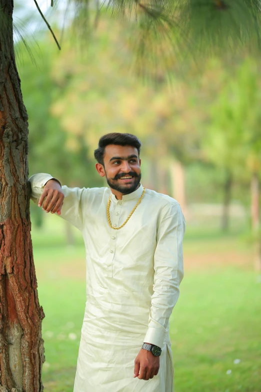 a man standing next to a tree in a park, a picture, hurufiyya, wearing a white shirt, elegant smiling pose, indian, profile image