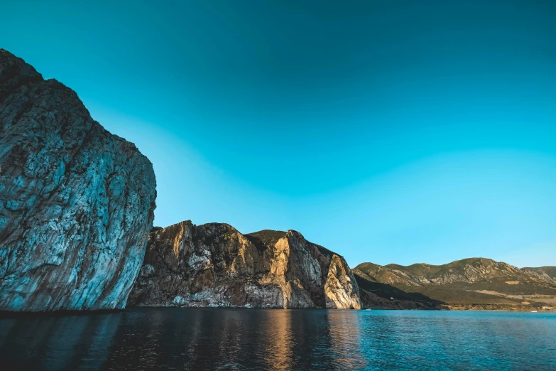 a large rock in the middle of a body of water, by Alexander Fedosav, pexels contest winner, romanticism, sharp cliffs, summer evening, clear blue skies, thumbnail