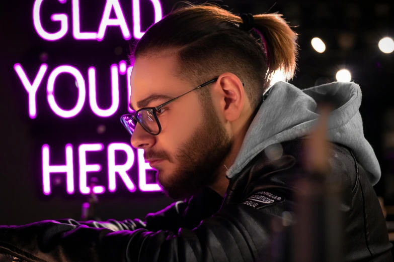 a man sitting in front of a neon sign that says glad you're here, by Julia Pishtar, trending on pexels, singer maluma, twitch streamer / gamer ludwig, purple and black, medium close-up shot