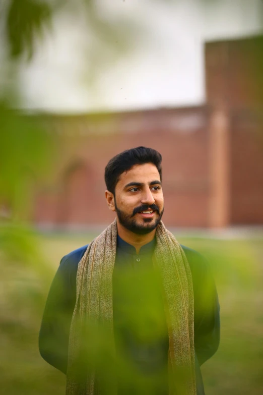 a man standing in front of a building wearing a scarf, pexels contest winner, bengal school of art, a portrait of rahul kohli, trees in background, slightly pixelated, profile image