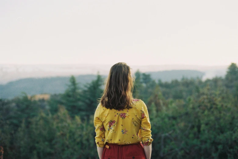 a woman standing on top of a lush green hillside, a picture, trending on unsplash, wearing yellow floral blouse, looking from behind, pine trees in the background, wearing red and yellow clothes