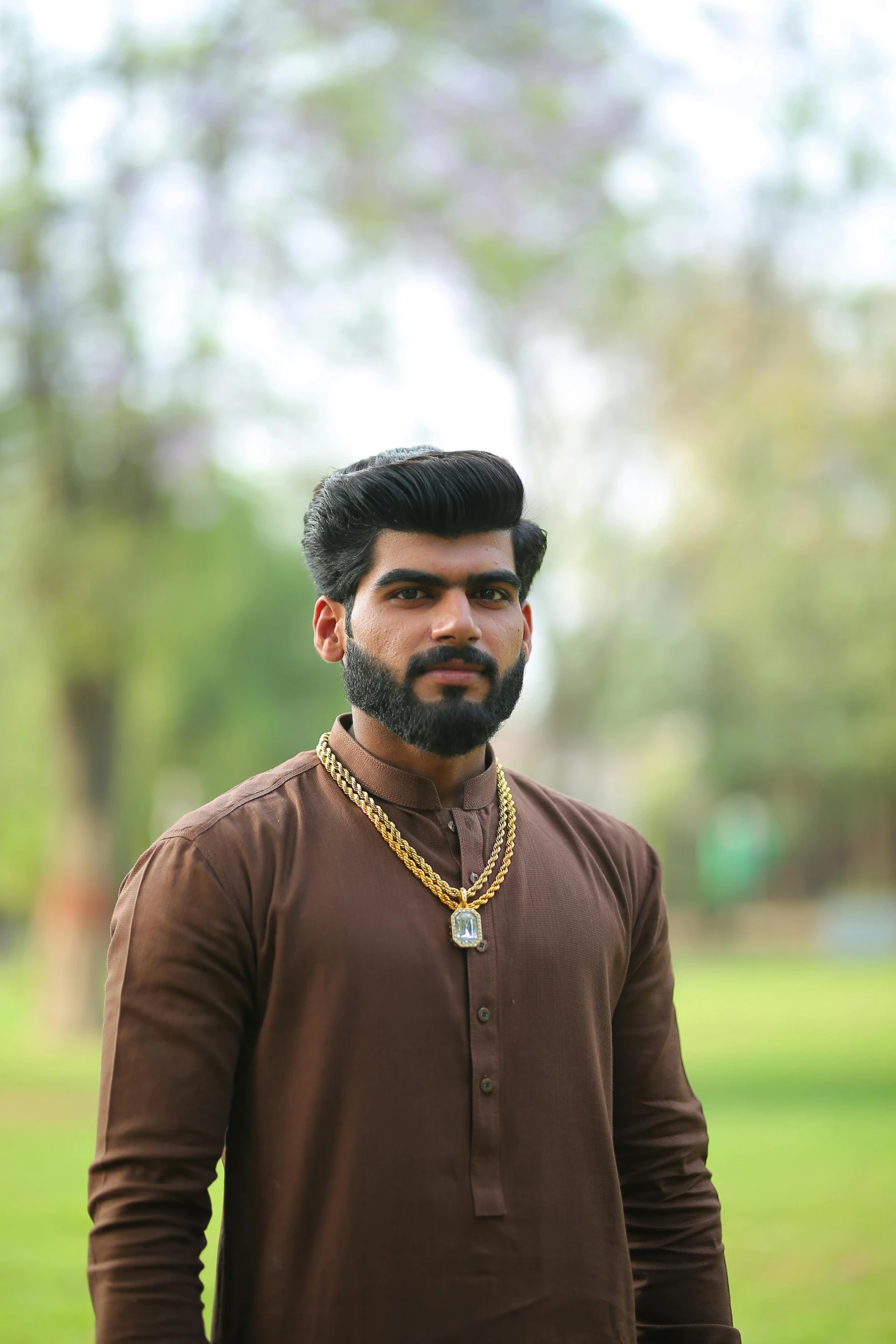 a man in a brown shirt standing in a park, by Max Dauthendey, pexels contest winner, hurufiyya, wearing gold jewellery, shaved beard, with kerala motifs, he is about 20 years old | short