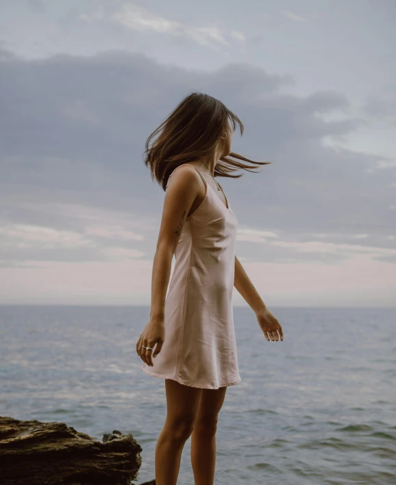 a woman standing on top of a rock next to the ocean, an album cover, unsplash, soft silk dress, outfit photo, short minidress, muted colours