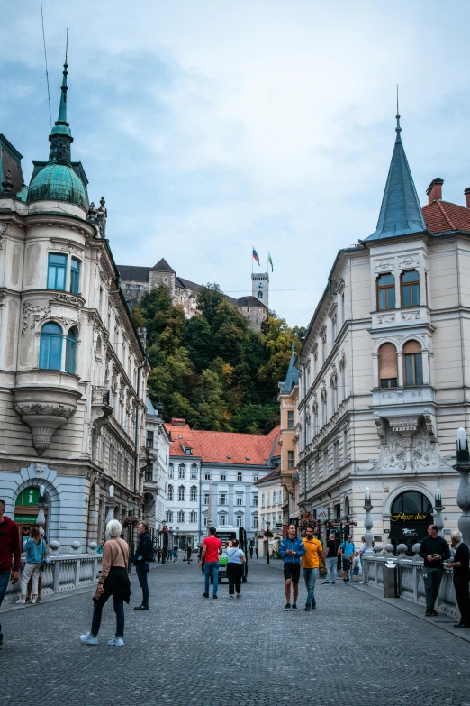 a group of people walking down a cobblestone street, a picture, trending on unsplash, baroque, castle on the mountain, square, slovenian, round buildings in background