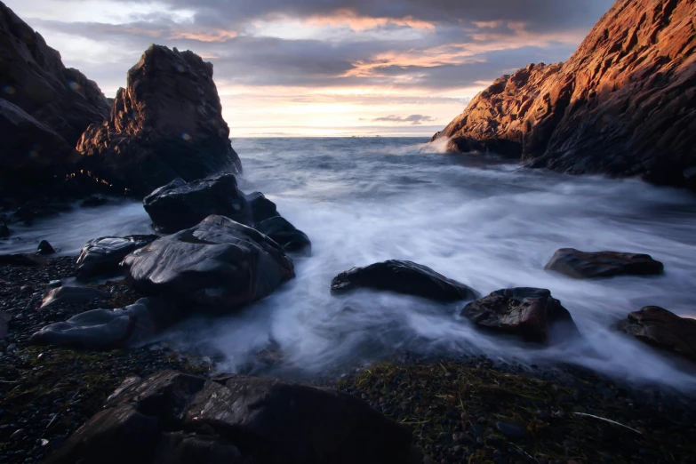 a group of rocks sitting on top of a rocky beach, by Andrew Geddes, unsplash contest winner, romanticism, waves crashing at rocks, cliff side at dusk, thumbnail, medium format. soft light