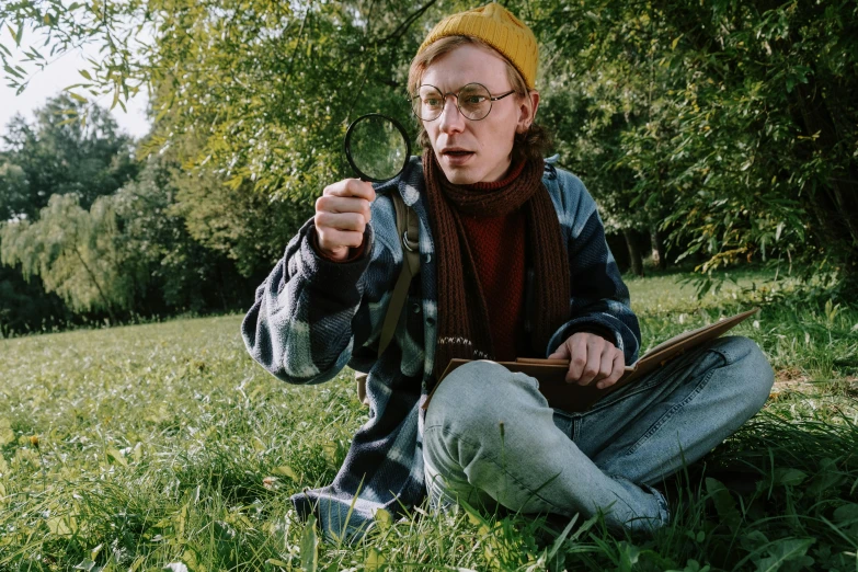 a man sitting in the grass pointing at something, inspired by Wes Anderson, pexels, hyperrealism, magnifying glass, eleanor tomlinson, square rimmed glasses, dressed as a scavenger