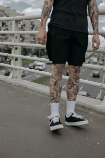 a man with tattoos standing on a bridge, a tattoo, inspired by Seb McKinnon, gray shorts and black socks, zoomed out full body, lowrider style, wearing black shorts