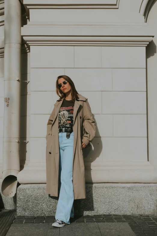 a woman standing in front of a building, light brown trenchcoat, graphic tees, jeans and t shirt, trending on vsco