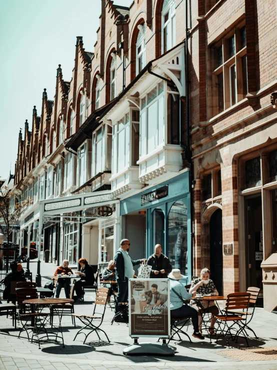 a group of people sitting outside of a building, pexels contest winner, arts and crafts movement, house's and shops and buildings, full daylight, barnet, breakfast