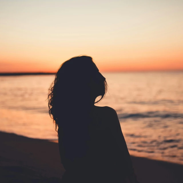 a woman standing on top of a beach next to the ocean, trending on pexels, romanticism, silhouette over sunset, girl with dark brown hair, woman's profile, sitting down