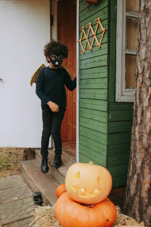 a person wearing a mask standing in front of a house, by Kristian Zahrtmann, pexels contest winner, winged boy, beistle halloween decor, with afro, leaning on door