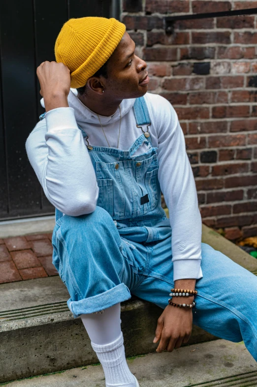 a man sitting on the steps of a brick building, an album cover, trending on pexels, wearing blue jean overalls, wearing a chocker and cute hat, looking from side!, non binary model